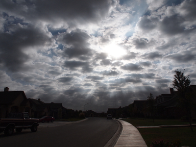 [The sun is at the upper center of the image with its rays flowing between the many clouds in the sky over what appears to be a dark neighborhood (dark because the clouds block most of the sun. The rays almost seem like petals of a flower radiating from the sun center.]